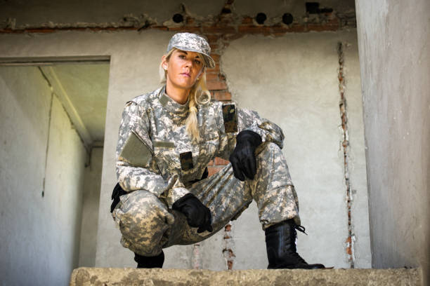 Female army soldier in military uniform is posing indoors on gray wall background.