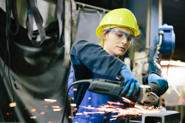 young female trainee welds steel with grinder in workshop, wearing work helmet and protective eyewear,