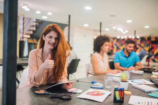 istock Joyful redhead woman using digital tablet and holding thumbs up while smiling at the office 1027777884