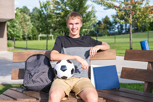 The image displays a male teenager sitting on a wooden bench in front of a school. On the bench are his backpack, school supplies and soccer ball.