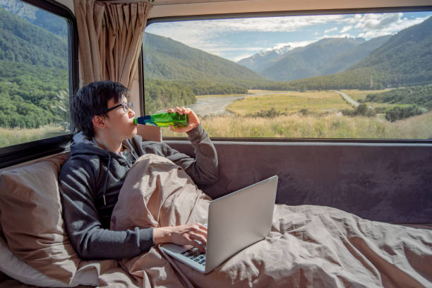 istock Young Asian man drinking beer and working with laptop computer on the bed in camper van with mountain scenic view through the window. digital nomad on road trip concept 1027797824