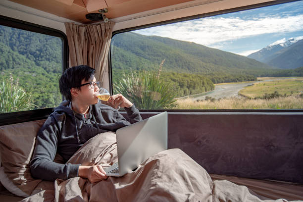 istock Young Asian man drinking beer and working with laptop computer on the bed in camper van with mountain scenic view through the window. digital nomad on road trip concept 1027797832