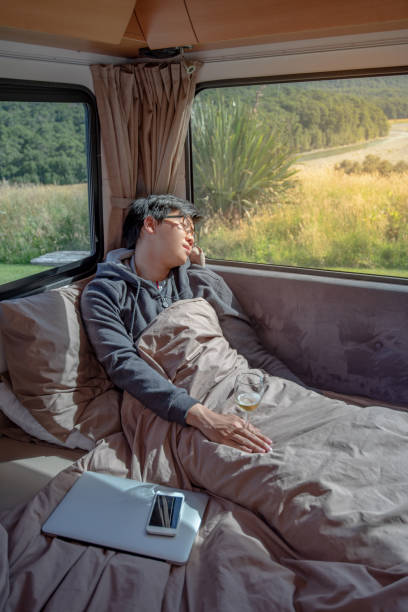 istock Young Asian man hangover after drinking beer in camper van with mountain scenic view through the window, laptop computer and smartphone on the bed. digital nomad on road trip concept 1027797852