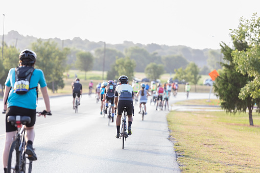 In this rear view, a large group of bike race for charity participants can be seen riding down an empty street.  There is a field and trees in the background.