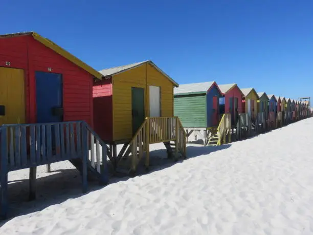 Red, yellow, blue and green beach huts stretching into the distance on wide sand under a deep blue sky