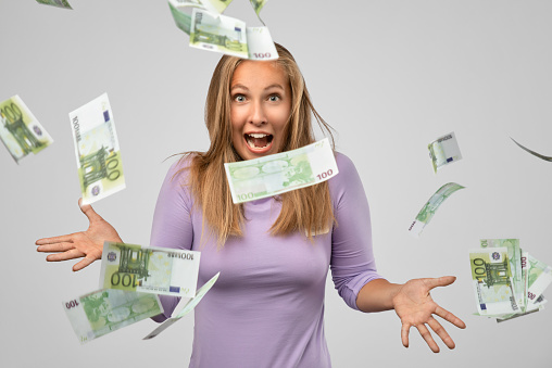 Portrait of cheerful glad blonde girl enjoying shower from 100 euro, flying money, gesturing with hands isolated on gray background