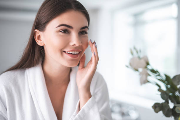 Satisfied girl doing face treatment in morning Portrait of pleased young lady touching cheek with hand. She putting cream on face skin indoor cheek photos stock pictures, royalty-free photos & images