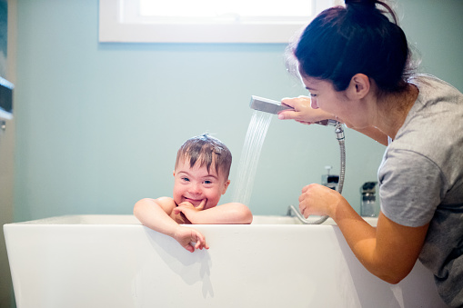 Down's syndrome sweet boy in the bath while Mom is washing him. He has blue eyes and blond hair. He is sitting in the bath and looking directly the camera. Mom watering her son with the shower. He as a cute face. The color and horizontal Photo was taken in Quebec Canada. There is copy space in this picture.