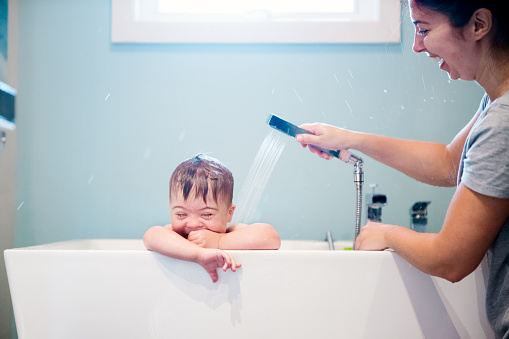 Down's syndrome sweet boy in the bath while Mom is washing him. He has blue eyes and blond hair. He is sitting in the bath and looking directly the camera. Mom watering her son with the shower. He as a cute face. The color and horizontal Photo was taken in Quebec Canada. There is copy space in this picture.