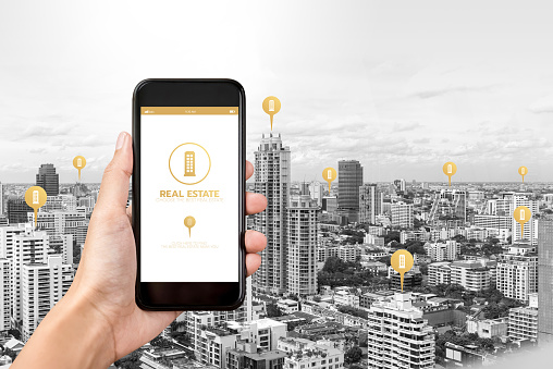 Hand holding smartphone with application to find  real estate on screen and buildings in the city as background