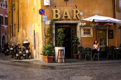 Rome, Italy: A young woman sits alone outside a vibrant yellow coffee bar smoking in Rome’s Trastevere neighborhood. Early morning shot.