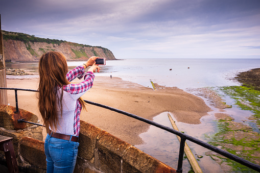 Robin Hood's Bay is an old fishing village on the Yorkshire coastline along England's East coast. Cliffs can be seen in the distance. The woman is traveling alone and she's enjoying the view of the beautiful destination. She's taking a photo with her mobile phone.