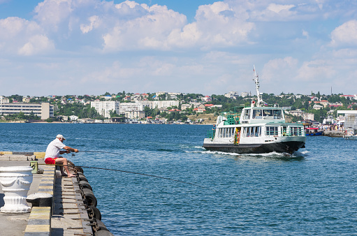 Russia, the peninsula of Crimea, the city of Sevastopol. 06/10/2018: Passenger ship in Sevastopol Bay and an angler with a fishing rod on the pier. View of the bay water area and the city