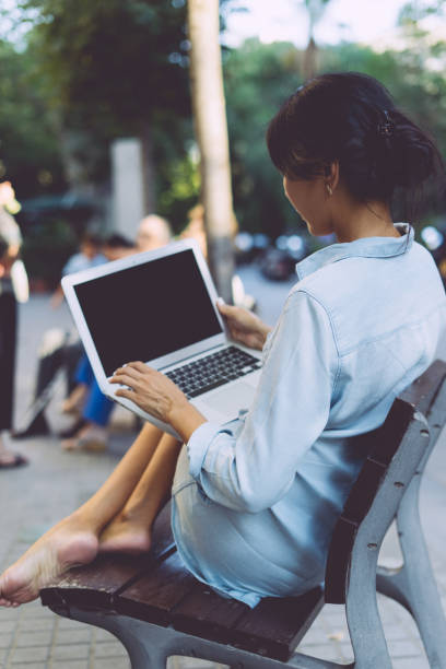 beautiful asian woman is sitting at city public space with a laptop connected to wi-fi. young web designer is working on a portable computer while sitting outdoors on a blurred urban background. - surfing wireless vertical outdoors lifestyles imagens e fotografias de stock