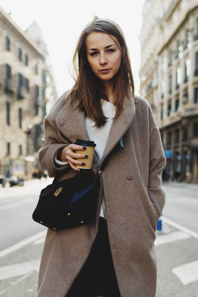 istock Portrait of successful student in a trendy oversized coat looking at the camera while walking outdoors in a city with coffee to go. Young business woman standing at the street on a sunny day. 1028091682