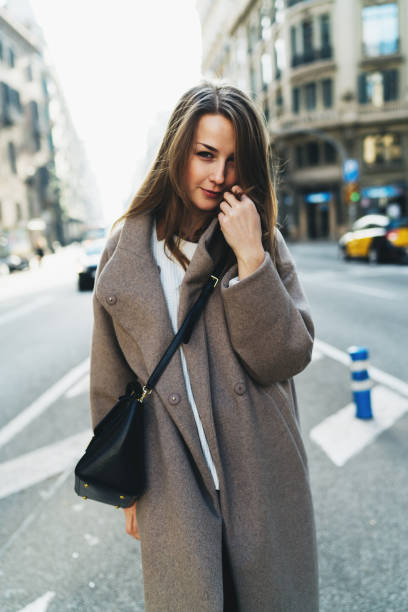 istock Shy student girl looking at the camera while standing on a blurred city background. Hipster girl with long hair wearing trendy oversized coat crossing the road on a warm sunny spring day. 1028091862