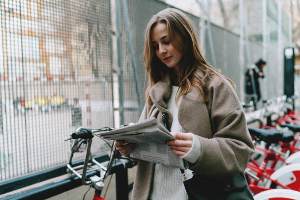 istock Young caucasian woman reading the interesting article in a newspaper while passing by the city bike rental station. Student girl in a trendy outfit standin with a daily magazine on urban background 1028091902