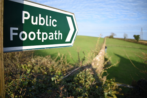 Closeup of a public footpath sign taken with a shallow depth of field, pointing across a green field with a person looking over a stone wall built through the field in Devon, UK.