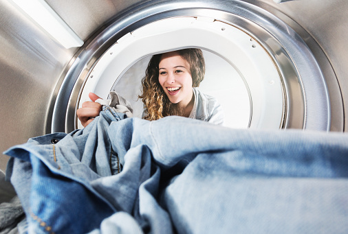 A lovely young woman with curly brown hair smiles as she reaches into an open tumble dryer  to remove a pair of jeans. Seen from inside the dryer.