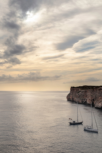High view of the sea and two boats in Menorca during the sunset in a overcast day, casting a strong orange color in the seascape