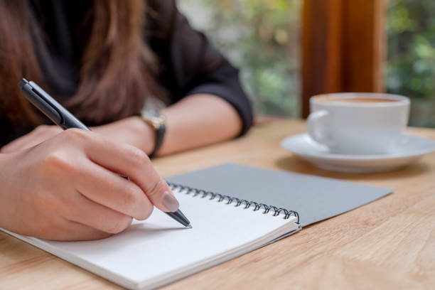 istock Closeup image of a woman's hand writing down on a white blank notebook with coffee cup on wooden table 1028142852