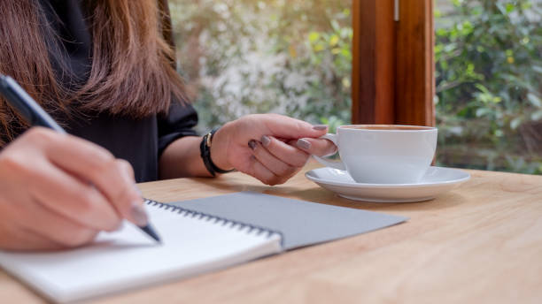 istock Closeup image of a woman's hand writing down on a white blank notebook while drinking coffee on wooden table 1028142888