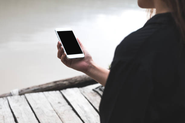 istock Mockup image of woman holding white mobile phone with blank black desktop screen while standing on the wooden pier 1028144116