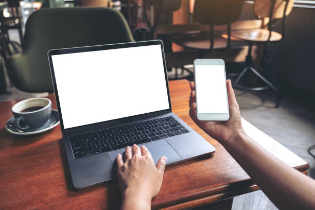 istock Mockup image of business woman holding mobile phone with blank white screen while using laptop on wooden table in cafe 1028147076