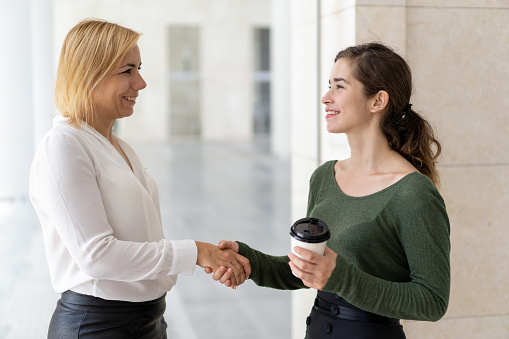 Friendly office colleagues shaking hands. Positive business women greeting each other, one of them drinking takeaway coffee. Meeting during coffee break concept