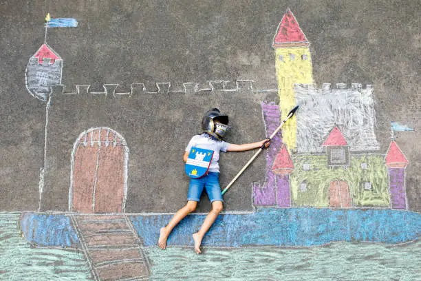Photo of Little active kid boy drawing knight castle and fortress with colorful chalks on asphalt. Happy child in helmet and with spear having fun with playing knight game and painting