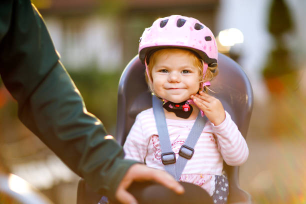 portrait of little toddler girl with security helmet on the head sitting in bike seat and her father or mother with bicycle. safe and child protection concept. family and weekend activity trip. - helmet bicycle little girls child imagens e fotografias de stock