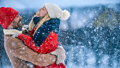 Handsome Young Couple Embrace and Happily Smile, Snow Falls on them. Couple in Love on the Backyard of Their Idyllic Home one Beautiful Winter Day.