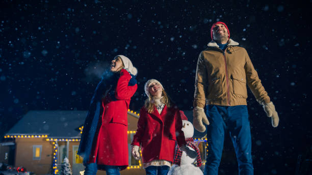 frohes neues jahrfeier, junge familie mit drei stehen im vorgarten blick in den himmel. am abend beim schnee ist fallen vater, mutter und niedliche kleine tochter schauen verwundert. haus für heiligabend mit girlanden geschmückt. - family house flash stock-fotos und bilder