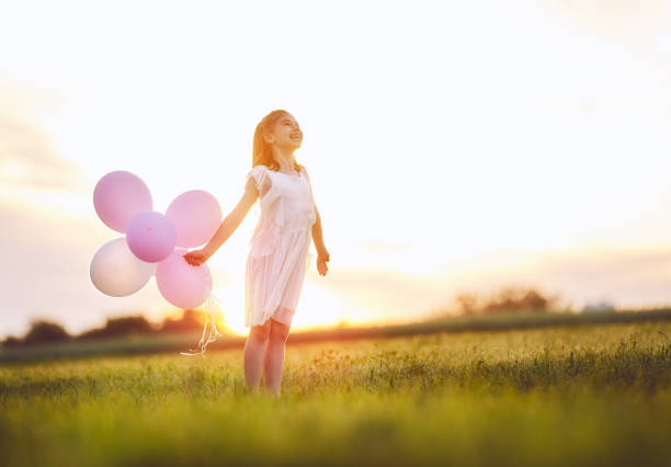 Happy kid is having fun on nature in the summer. Child is laughing and playing on meadow at sunset background. Girl with air balloons.