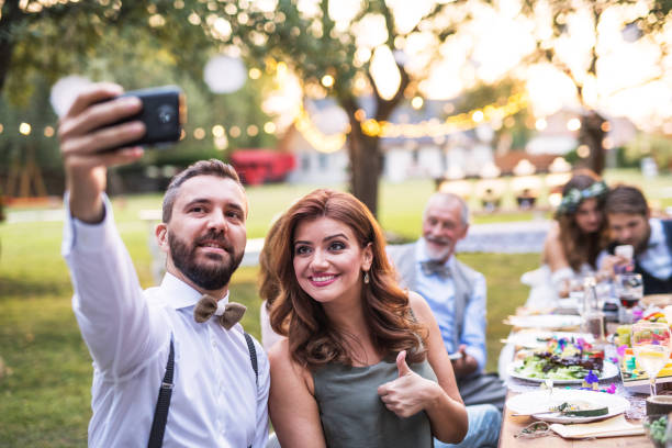 A couple taking selfie at the wedding reception outside in the backyard. A couple taking selfie at the wedding reception outside in the backyard. Bride, groom and guests sitting at the table. guest photos stock pictures, royalty-free photos & images