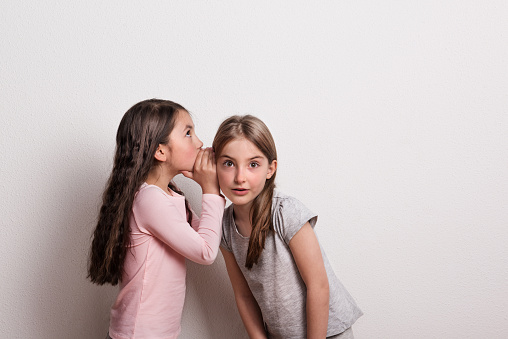 Two small cheerful girls standing in a studio. A small girl whispering something in an ear of her friend.