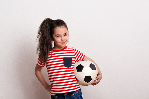 A small girl holding a soccer ball and striped T-shirt standing in a studio.