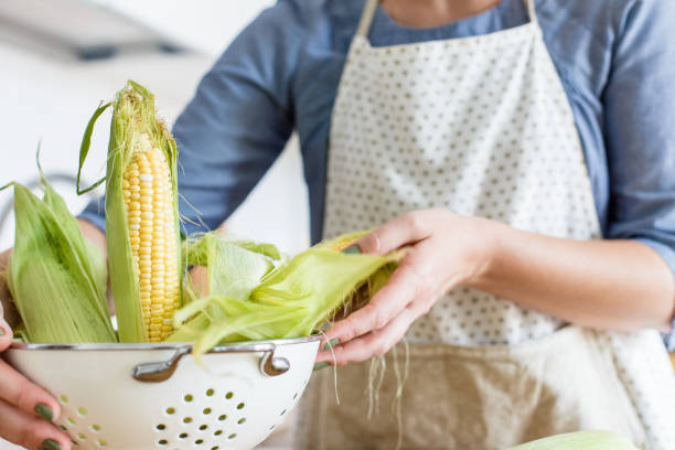 istock Woman holding colander with yellow corn cobs. View on white modern scandinavian style kitchen 1028237472