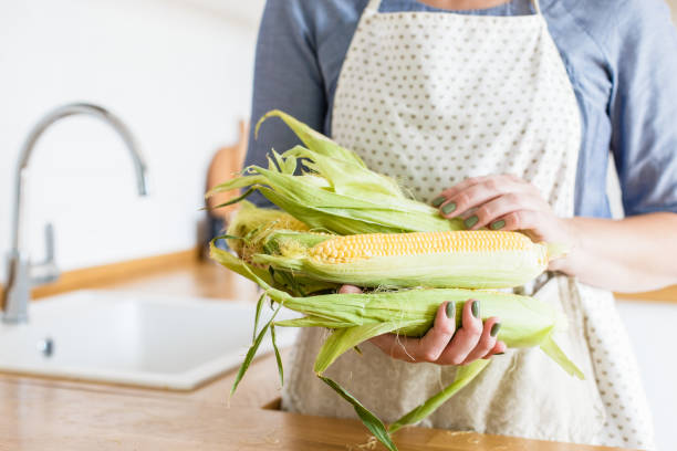 istock Woman holding colander with yellow corn cobs. View on white modern scandinavian style kitchen 1028237578
