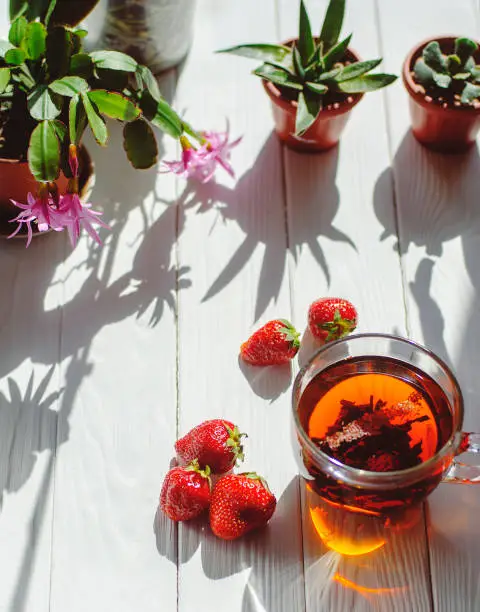 Photo of Close-up image of glass cup of tea, red strawberries, flowers in pots on light wooden background.