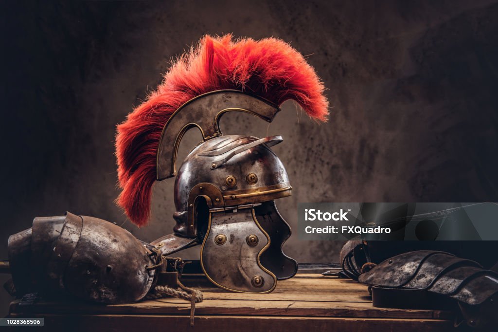Complete combat equipment of the ancient Greek warrior lie on a box of wooden boards. Complete combat equipment of the ancient Greek warrior lie on a box of wooden boards. Isolated on a dark background. Roman Stock Photo
