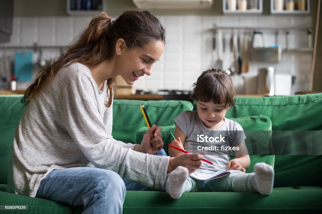 Mom and kid girl drawing with colored pencils at home Smiling baby sitter and preschool kid girl drawing with colored pencils sitting on sofa together, single mother and child daughter playing having fun, creative family activities at home concept Nanny Stock Photo