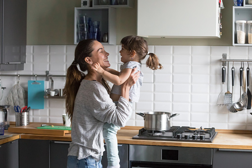Smiling loving single mother holding cute little child daughter having fun together, happy family of young caring mom and excited kid girl laughing playing at home while cooking in the kitchen