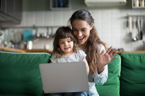 Happy mother and kid daughter waving hands looking at web camera using laptop for video call, smiling mom and child girl having fun greeting online by computer webcam making videocall via application