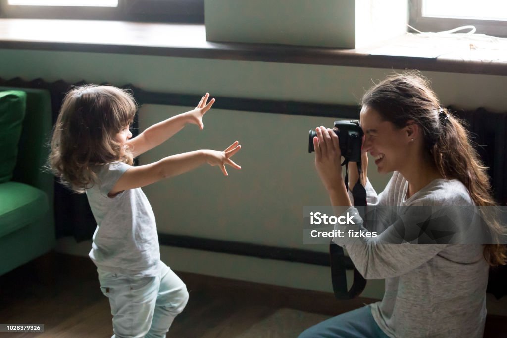 Mom photographer making photo of kid daughter on digital camera Mom photographer having fun playing making photo of artistic cute kid daughter at home, young mother photographing little child girl or shooting video on digital camera, funny family photo session Home Video Camera Stock Photo