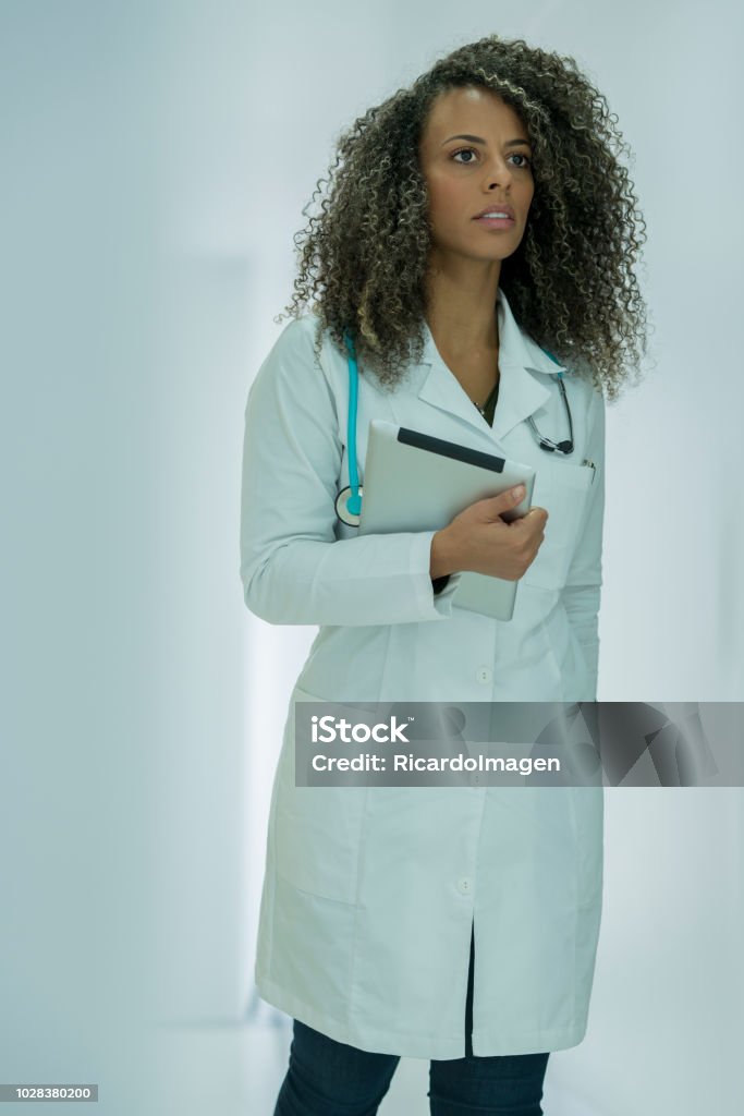 Female doctor walking through the hospital Doctor of Latin race, between 30 and 35 years old, walking in an attitude of concern for the hospital corridor, with a tablet in her hand and the stethoscope hanging from her neck Adult Stock Photo