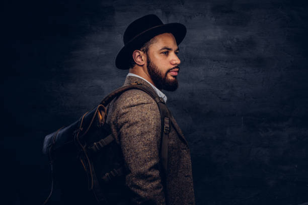 istock Handsome Afro-American traveler in a brown jacket and hat with the backpack, stands in a studio. 1028398616