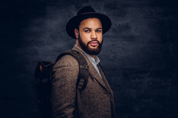 istock Handsome Afro-American traveler in a brown jacket and hat with the backpack, stands in a studio. 1028401156