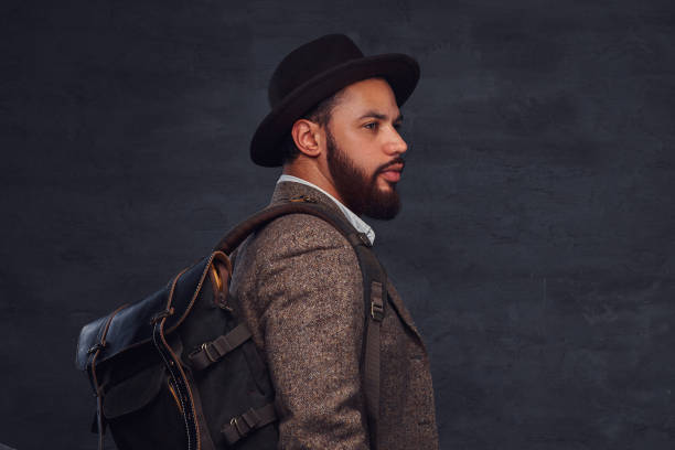 istock Handsome Afro-American traveler in a brown jacket and hat with the backpack, stands in a studio. 1028401190