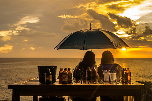 Negril, Jamaica - May 30 2015: Two caucasian women sitting at table with Red Stripe Beer holding an umbrella looking at the sunset in Negril, Jamaica.